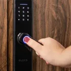 Mão com unha vermelha usando fechadura digital preta com biometria azul em porta de madeira.