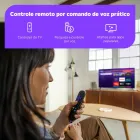 Mulher segurando controle remoto preto Roku Controle Voz com TV ligada.